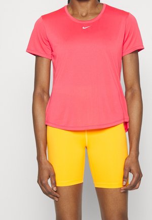 Femme portant un t-shirt Nike rose vif à manches courtes et un short de vélo jaune, debout devant un fond clair uni.