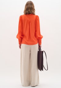 Orange blouse with puff sleeves, textured finish, back pleat detail; paired with wide-leg cream pants and a brown handbag.
