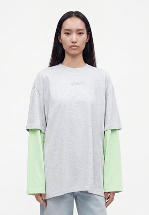 Jeune femme aux longs cheveux foncés porte un t-shirt gris clair à manches courtes par-dessus un t-shirt vert pâle à manches longues, associé à un jean bleu clair.