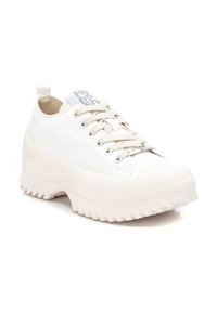 Zapatilla blanca con parte superior de lona lisa, cordones beige y una suela de goma gruesa y texturizada con estrías. Presenta una etiqueta de logo en la lengüeta.