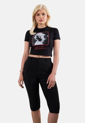 Femme blonde portant un t-shirt graphique noir court avec un imprimé Marilyn Monroe et du texte, associé à un pantalon noir ajusté longueur genou.