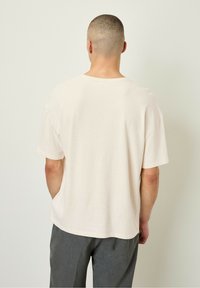 Písková bavlněná trička s volným střihem, krátkými rukávy a kulatým výstřihem. Má hladkou texturu a neformální design.