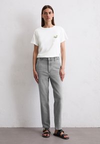 T-shirt en coton blanc avec un petit graphique vert, associé à un jean gris clair à coupe droite et poches classiques, et des sandales noires.