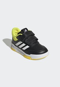 Zapatilla deportiva de cuero negro con tres franjas blancas, correas de Velcro, un collar acolchado amarillo y una suela de goma blanca y amarilla.