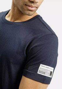 Granatowa bawełniana koszulka z okrągłym dekoltem, z metką na lewym rękawie z napisem "CROSS JEANS" oraz instrukcjami pielęgnacyjnymi.