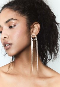 Lange guldsmykker med cirkulære toppe og faldende teksturerede strenge, hængende elegant nær modelens ansigt mod en neutral baggrund.