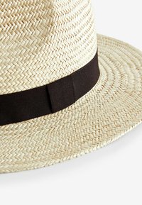 Chapeau fedora en paille avec une texture tissée beige clair et une bande en tissu marron foncé autour de la base.