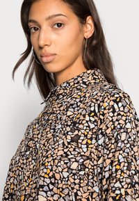 Blusa preta com um padrão geométrico em pedras nas cores bege, azul claro e laranja. Projetada com colarinho e fecho de botão.