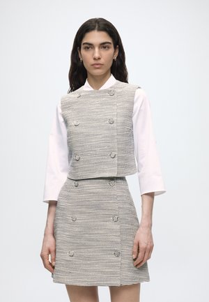 Kvinde med langt mørkt hår iført en hvid langærmet skjorte under en gråstripet ærmeløs vest og matchende nederdel med knapdetaljer.