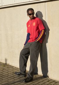 Κόκκινη φανέλα με κίτρινο λογότυπο της Adidas και έμβλημα της Ισπανίας πάνω από ναυτικές μακριές μανίκες με κόκκινες και κίτρινες ρίγες; γκρίζα τζιν και μαύρα παπούτσια.