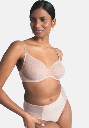 Frau trägt einen hautfarbenen Spitzen-BH und passende High-Waist-Slips, steht mit einer Hand in der Hüfte, lächelt und blickt zur Seite.