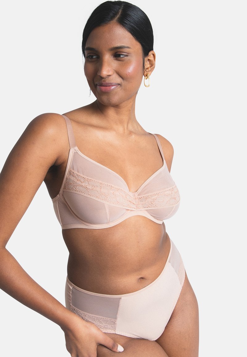 Frau trägt einen hautfarbenen Spitzen-BH und passende High-Waist-Slips, steht mit einer Hand in der Hüfte, lächelt und blickt zur Seite.