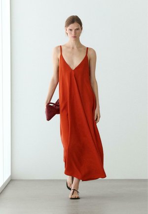 Kobieta spacerująca w pomieszczeniu, ubrana w luźną czerwoną, bezrękawną sukienkę maxi, czarne sandały i trzymająca małą plecioną czerwoną torebkę.