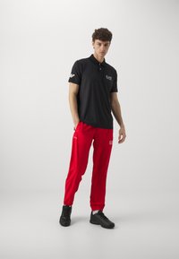 Schwarzes Poloshirt mit kurzen Ärmeln, mit einem Logo auf der Brust; kombiniert mit leuchtend roten Jogginghosen und schwarzen Sportschuhen.
