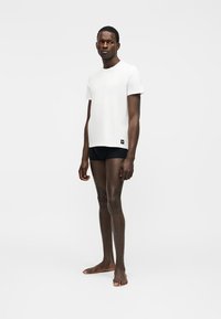 Uomo in piedi a piedi nudi, indossa una maglietta bianca e pantaloncini neri su sfondo bianco semplice.