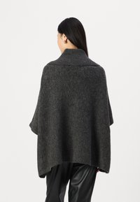 Poncho tricoté gris foncé avec un col haut, une forme ample et une texture douce, atteignant les hanches, porté sur un pantalon noir.