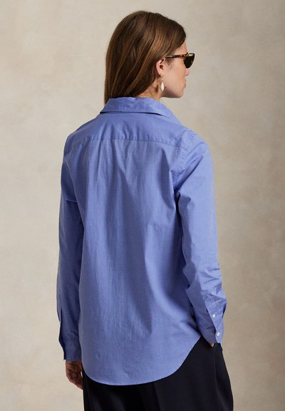 Kvinne med brunt hår som har på seg skilpaddemønstrede solbriller og en blå skjorte med lange ermer, vist bakfra mot en nøytral bakgrunn.