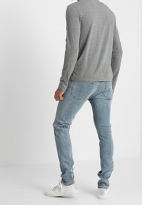 Maglietta grigia a maniche lunghe indossata con jeans in denim azzurri e sneakers bianche. L'outfit presenta una vestibilità casual e un design minimalista.