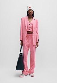 Blazer rosa su misura e pantaloni larghi abbinati, indossati con un top corto, un berretto bianco strutturato e una borsa a tracolla nera. Sneakers chiare.