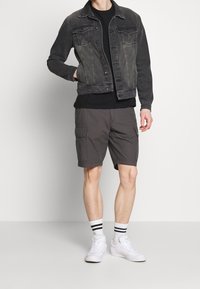 Grijze denimjas, zwart t-shirt, grijze cargoshorts, witte sneakers met zwartaccent sokken. Model staat in een neutrale omgeving.