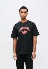 Melna kokvilnas T-krekls ar sarkanu "ADIDAS" uzrakstu un trīs svītru logotipu. Īsas piedurknes un apaļa kakla izgriezums. Vienkāršs dizains.