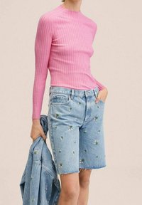 Różowa, prążkowana bluzka z długim rękawem w połączeniu z jasnoniebieskimi szortami dżinsowymi, zdobionymi haftem kwiatowym. Model trzyma dżinsową kurtkę.