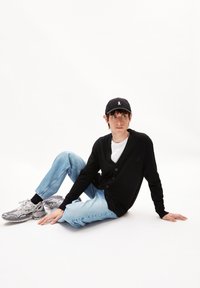 Schwarze Strickjacke mit V-Ausschnitt und Knöpfen, kombiniert mit hellblauen Jeans. Graue Sneakers mit detaillierten Mustern und einer schwarzen Mütze.