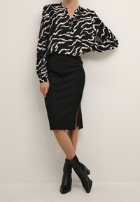 Femme portant un chemisier imprimé zèbre noir et blanc, une jupe crayon noire mi-genou avec une fente, et des bottines à talons noires, debout contre un mur uni.