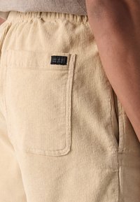 Pantalons en velours côtelé de couleur jaune pâle, avec une surface texturée, une taille élastique et une poche arrière avec une étiquette noire.