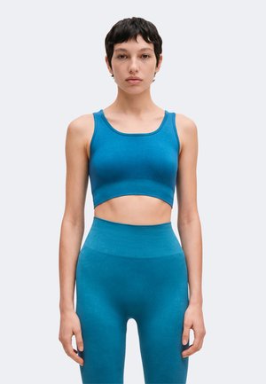 Mujer de pelo corto y oscuro lleva un top corto azul sin mangas y unos leggings de tiro alto a juego, de pie contra un fondo claro y liso.