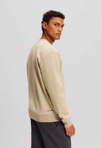 Sudadera beige de tejido suave con mangas largas, cuello redondo y dobladillo acanalado; presenta un pequeño logo de la marca en la cintura.
