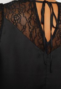 Vêtement noir avec détail floral en dentelle et tissu en mesh près de l'encolure, noué avec de fines cordelettes noires à l'arrière.