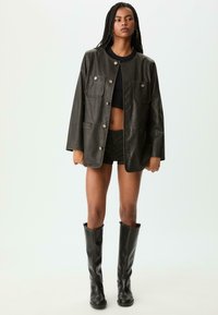 Sort læderjakke med rund krave, frontlukning med knapper, to brystlommer, båret over en sort crop top og stregede shorts.