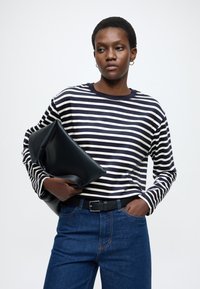 Tricou cu mânecă lungă în dungi bleumarin și alb, blugi de culoare albastru închis, ținând o geantă plic din piele neagră, având o curea neagră cu cataramă argintie.