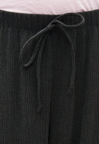 Μαύροι υφασμάτινοι παντελόνια με υφή και κορδόνι δεμένο σε φιόγκο στη λαστιχένια μέση, φορεμένοι με ένα ανοιχτό ροζ κορμάκι που έχει μέσα στην μέση.