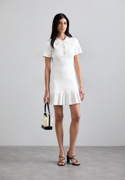 Witte polojurk met korte mouwen en een verlaagde taille met een geplooide zoom. Model houdt een kleine zwart en crèmekleurige handtas vast en draagt zwarte hakken.