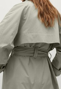 Oliven grøn trench coat med en struktureret design, brede ærmer og taljebælte. Stoffet ser glat ud med minimale synlige sømme.