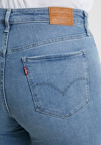 Jeans de mezclilla azul claro con un bolsillo trasero visible que presenta un diseño de costura curva y una etiqueta de cuero marrón. La tela tiene una textura suave.