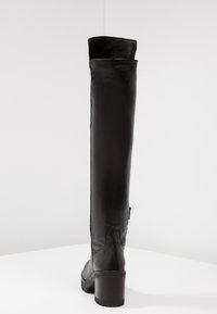 Bota negra por encima de la rodilla, hecha de cuero liso, con tacón grueso, diseño curvado y ligero acolchado en la parte superior.