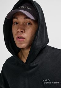 Czarna bluza z kapturem o teksturowanym wykończeniu z białym nadrukowanym tekstem na klatce piersiowej. Szara czapka z haftem "LEAVE NO TRAIL" na froncie.