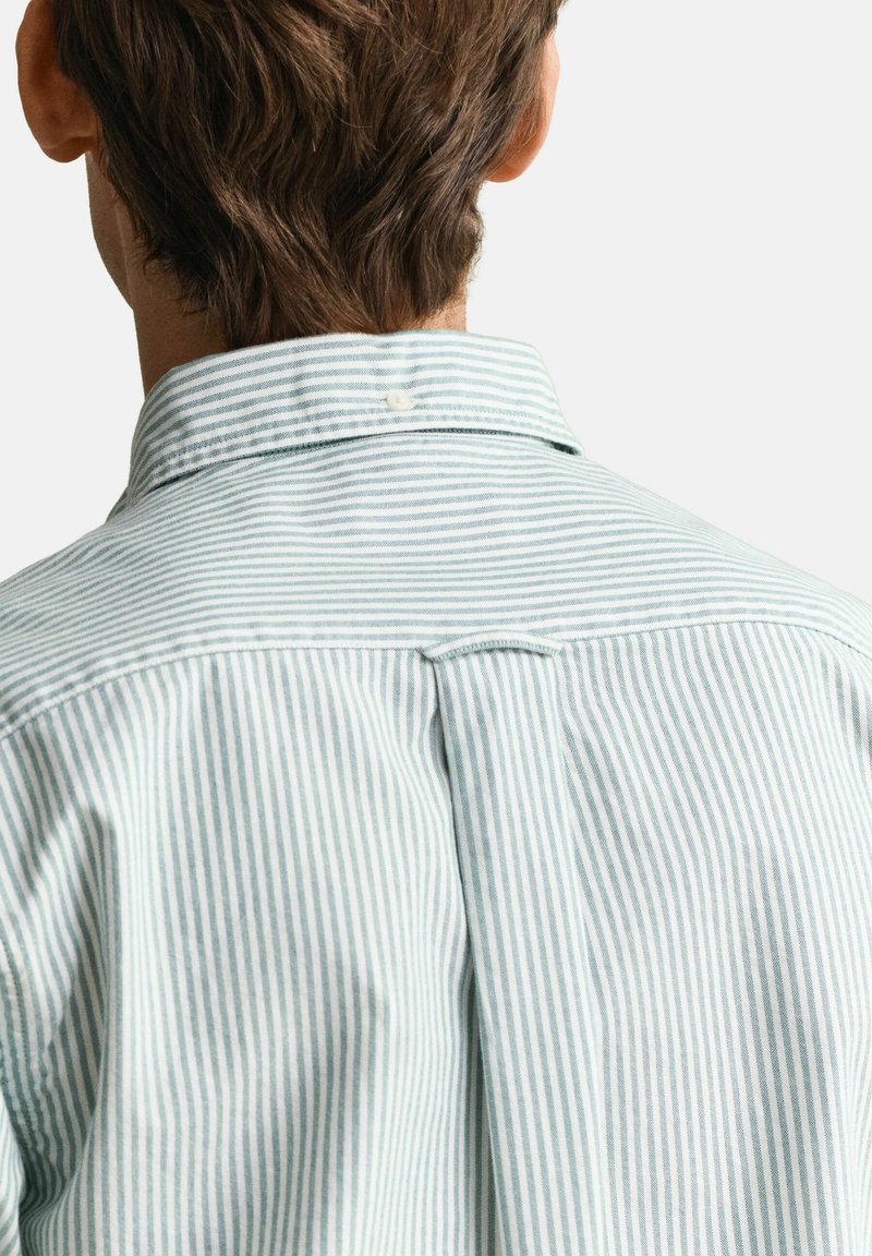 Uomo con capelli castani corti che indossa una camicia a righe verticali bianche e azzurre, vista posteriore, sfondo chiaro uniforme.