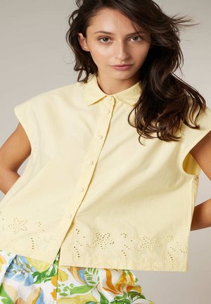 Femme aux longs cheveux bruns portant un chemisier sans manches jaune pâle à boutons et un pantalon fleuri multicolore, posant avec les mains sur les hanches.