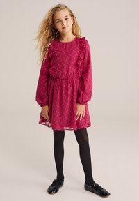 Mlada deklica z dolgimi blond lasmi, oblečena v teksturirano magentasto obleko z volančki, črne nogavice in črne Mary Jane čevlje, stoji na nevtralni podlagi.