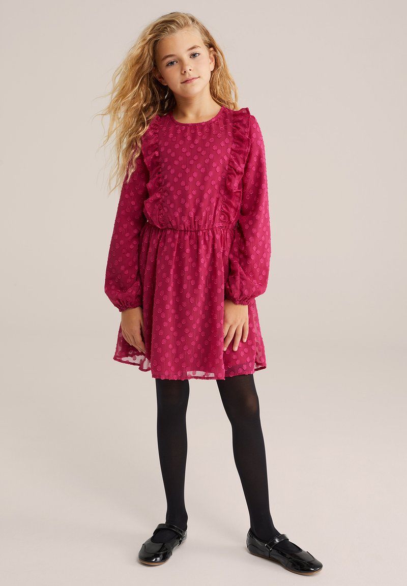 Mlada deklica z dolgimi blond lasmi, oblečena v teksturirano magentasto obleko z volančki, črne nogavice in črne Mary Jane čevlje, stoji na nevtralni podlagi.