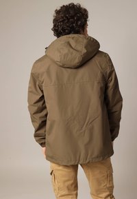 Bruine hooded jas van waterafstotend materiaal met elastische manchetten en een eenvoudig ontwerp. Gecombineerd met kaki cargobroek.