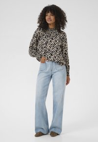 Blouse noire et beige à motifs avec un col froncé, associée à un jean large bleu clair. Le tissu semble doux et texturé.