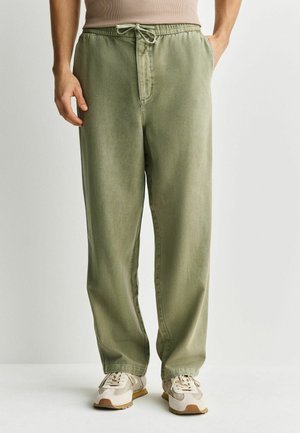 Man wearing olive green drawstring pants and beige sneakers, standing against a plain light background.