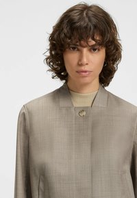 Beige, strukturierte Jacke mit Stehkragen, einem einzigen Knopfverschluss und geraden Ärmeln. Der Stoff hat einen dezenten Glanz.