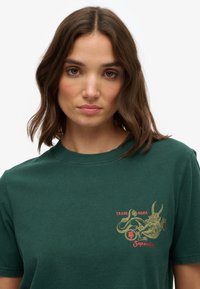Grøn bomulds t-shirt, kortærmet, med et grafisk design i guld og rød, der viser en skabning og teksten "Superdry" på venstre bryst.