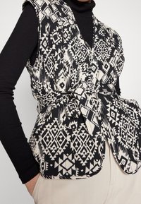 Svartvit mönstrad väst med midjeband, prydd med geometriska mönster och en lös, ärmlös passform. Bärs över en svart polotröja.